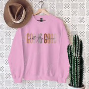 Pink Christian clothing women sweatshirt with God is Good design hanging on wall with brown hat and cactus