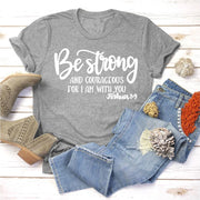 Gray t-shirt with Be Strong Joshua 1:9 quote paired with blue jeans and tan boots faith based apparel