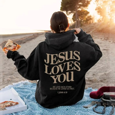 Person wearing black faith-based hoodies with Jesus Loves You message sitting on beach picnic