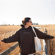 Woman wearing dark faith-based hoodies with Jesus Loves You text standing on wooden bridge in field