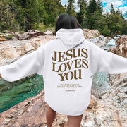 White faith-based hoodies with Jesus Loves You graphic and scripture 1 John 4:19 worn outdoors by rock river