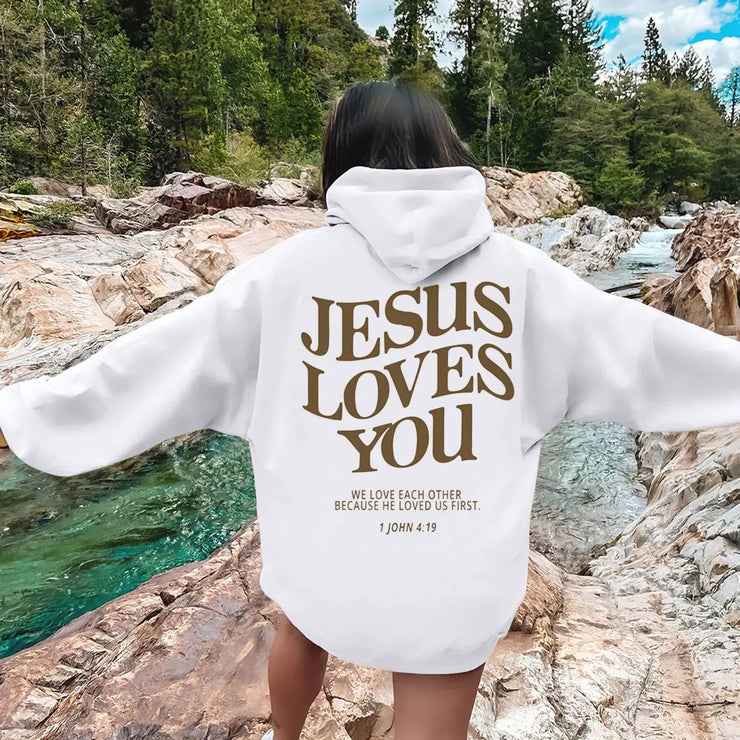 White faith-based hoodies with Jesus Loves You graphic and scripture 1 John 4:19 worn outdoors by rock river